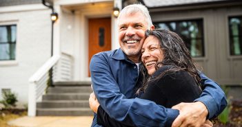 Two people hug with bright smiles in front of a house.