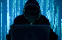 Man in a black hoodie behind a computer screen.