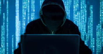 Man in a black hoodie behind a computer screen.